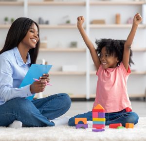 Emotional little black girl with bushy curly hair raising hands up, sitting on floor with friendly pretty african american woman, playing with colorful wood blocks, having session with psychologist